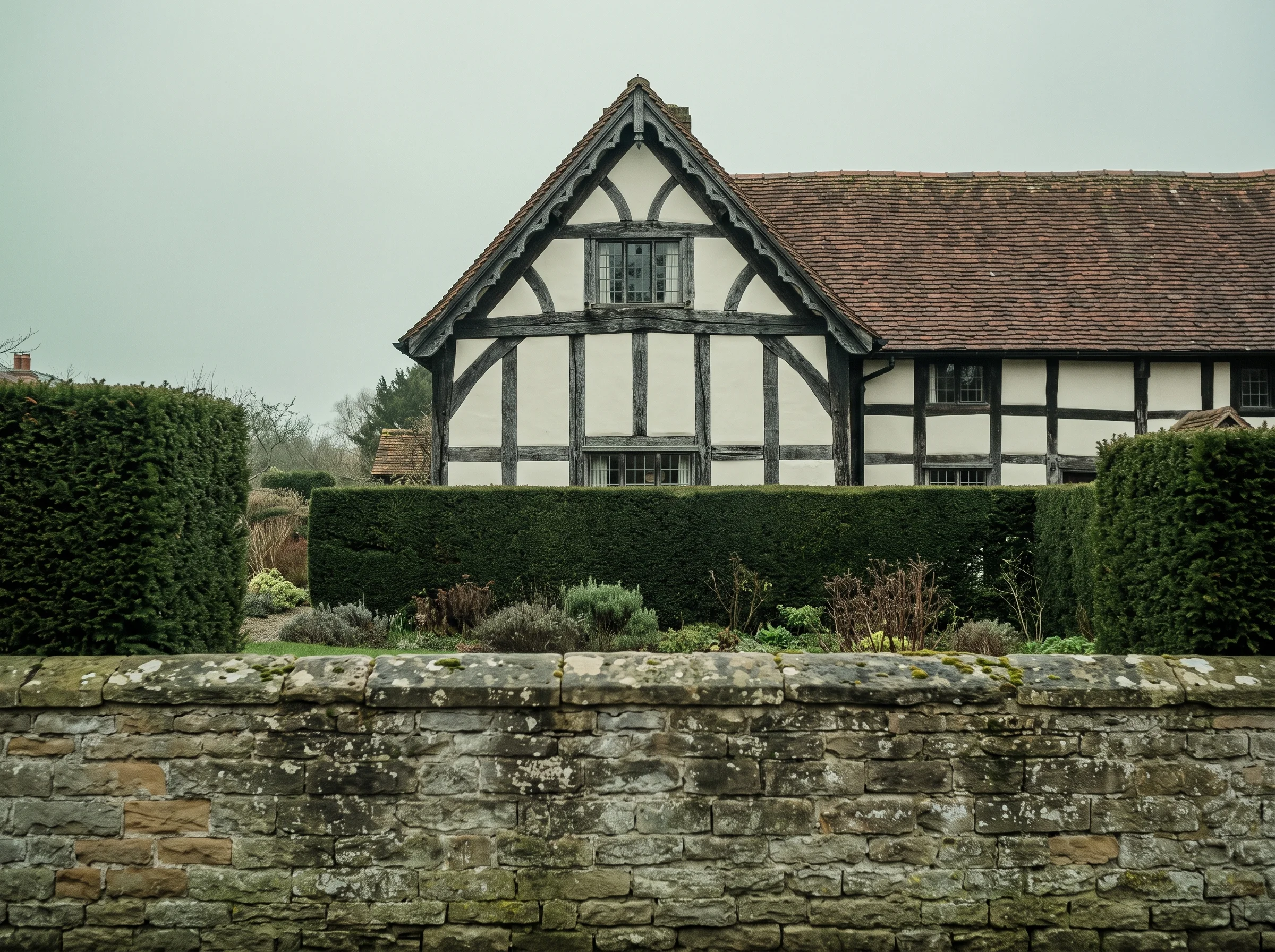 A timber-framed gable seen over a clipped hedge, stone wall in the foreground